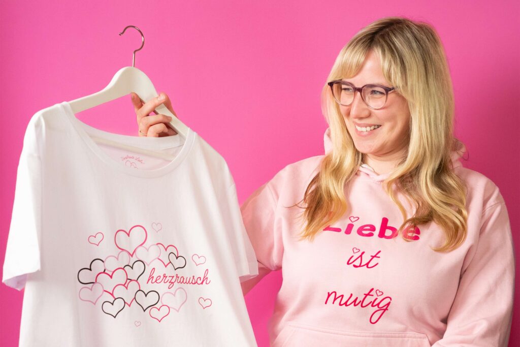 Eine Frau schaut lächelnd auf das Herzrausch T-Shirt, welches sie in der Hand hält.